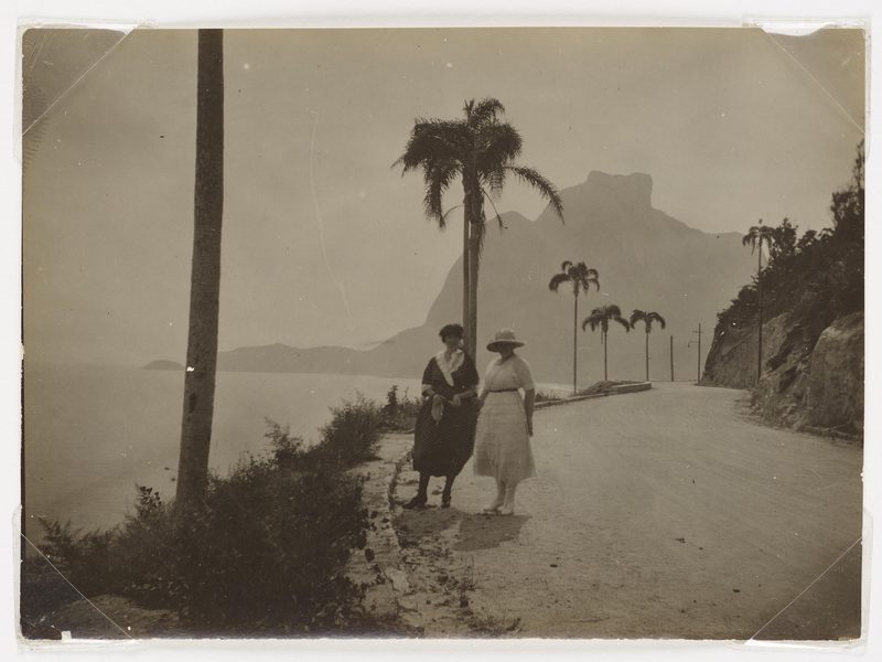 Two women walking along the road by the sea under palm trees, early 1900s